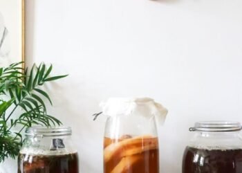 Glass jars with infused liqueurs with ingredients placed on wooden table near green plant near wall with decorations at home