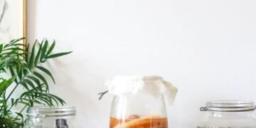 Glass jars with infused liqueurs with ingredients placed on wooden table near green plant near wall with decorations at home