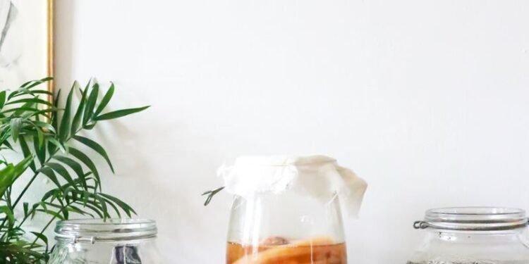 Glass jars with infused liqueurs with ingredients placed on wooden table near green plant near wall with decorations at home